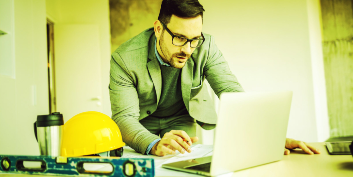 Dedicated hardworking architect standing in building in construction process and using laptop for improving blueprints.