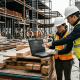A project manager and a site colleague reviewing job cost data on a laptop at an active commercial construction site.